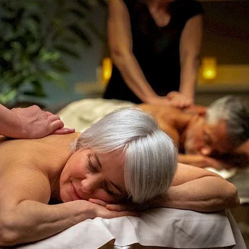 Two clients side by side on massage tables, draped, with therapists working on each